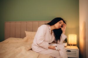 Frustrated woman sitting up in bed at night, wiping sweat from forehead with tangled sheets around her legs - alt="menopausal woman experiencing night sweats