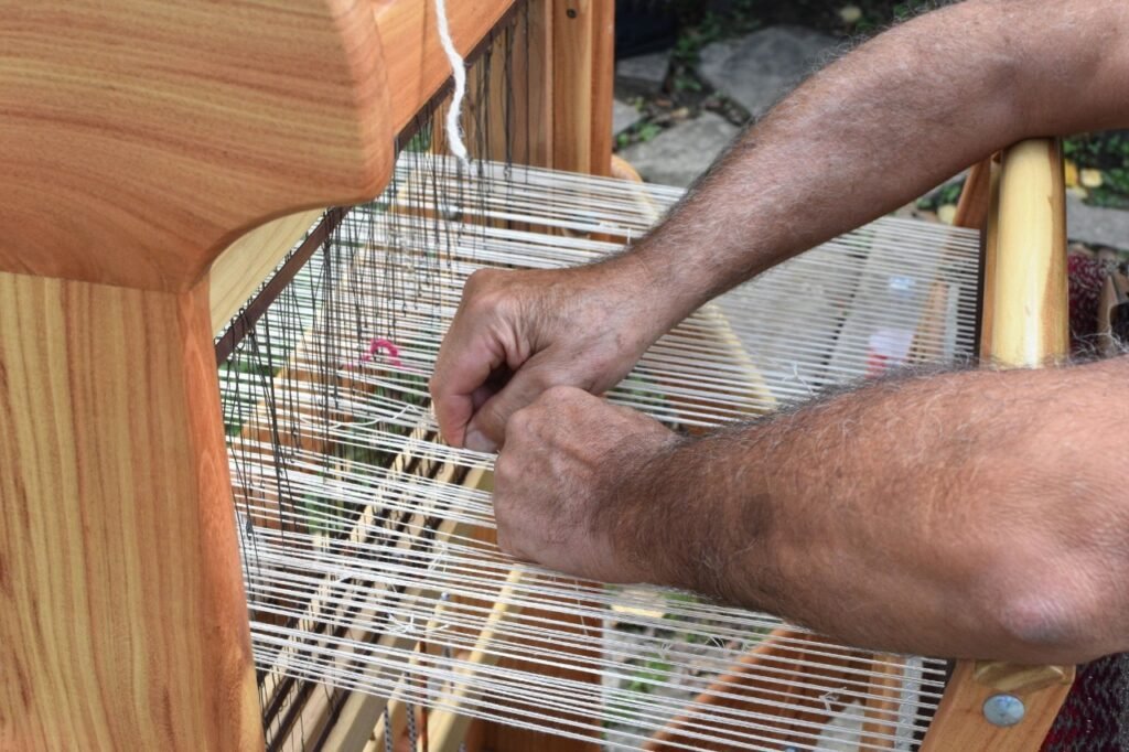 Carlos' hands guiding thread through his 1897 loom, with a shallow focus that blurs the modern world outside the workshop window.