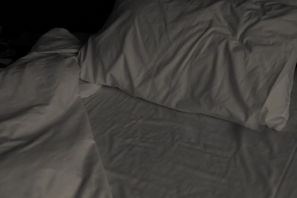 A moody, artistic shot of a disheveled bed in the middle of the night, lit only by the faint blue glow of a smartphone screen on a nightstand. The sheets are rumpled and tangled