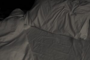 A moody, artistic shot of a disheveled bed in the middle of the night, lit only by the faint blue glow of a smartphone screen on a nightstand. The sheets are rumpled and tangled